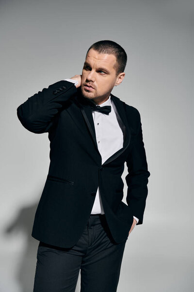 Человек в черном смокинге стоит на сером фоне, настраивая воротник..