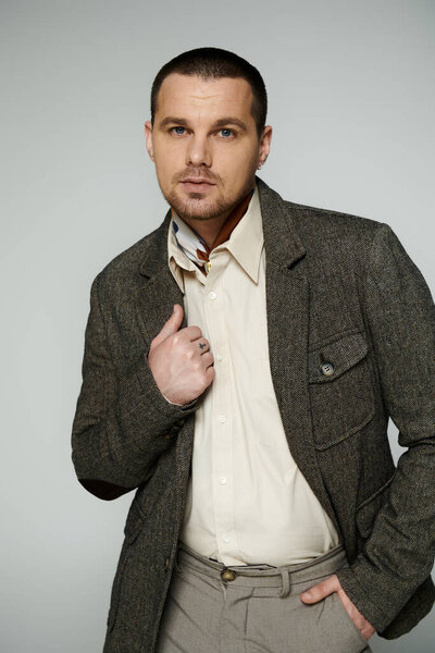 Man in tweed jacket, white shirt, and scarf poses with hand in pocket.