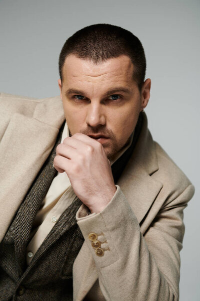 A stylish man in a beige coat, with a thoughtful expression, gazes off into the distance.