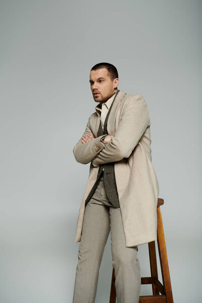 A stylish man in a beige coat stands with his arms crossed, leaning against a wooden stool.
