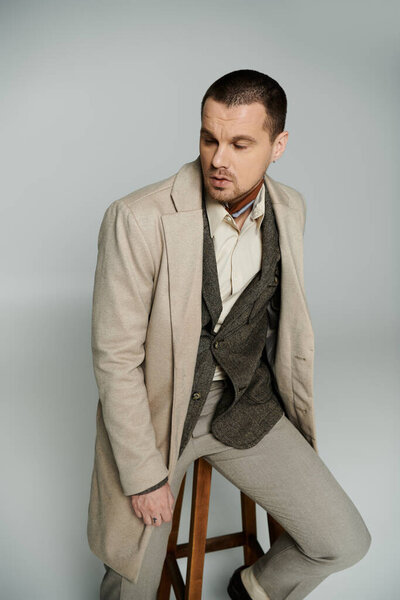 A stylish man in a long beige coat sits on a stool.