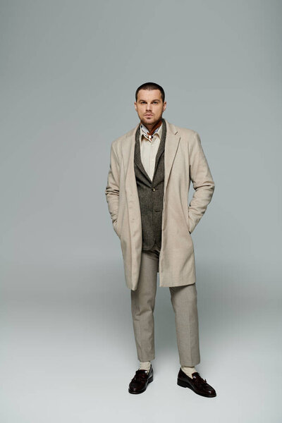 Человек в бежевом пальто и брюках, коричневые мокасины, стоит на белом фоне.