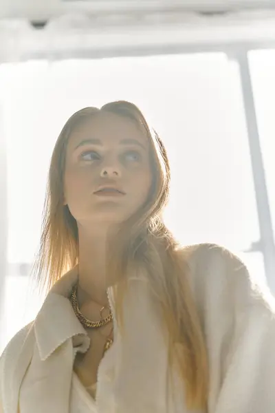 stock image A young woman in elegant attire looks out a window, bathed in soft sunlight.