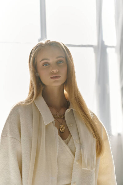 A young woman, wearing a stylish white outfit, stands in a sunlit room.