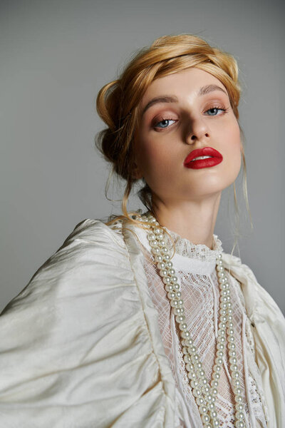 A young woman in a white lace blouse with blonde hair, red lipstick, and a pearl necklace poses.