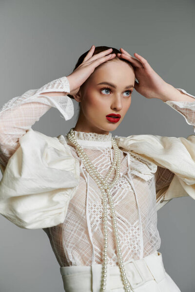 A young woman in a white lace blouse with pearl necklace, her hands framing her face.