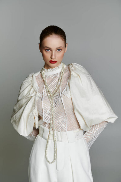 Woman in white lace top, pants, with pearl necklace, gazes intently.