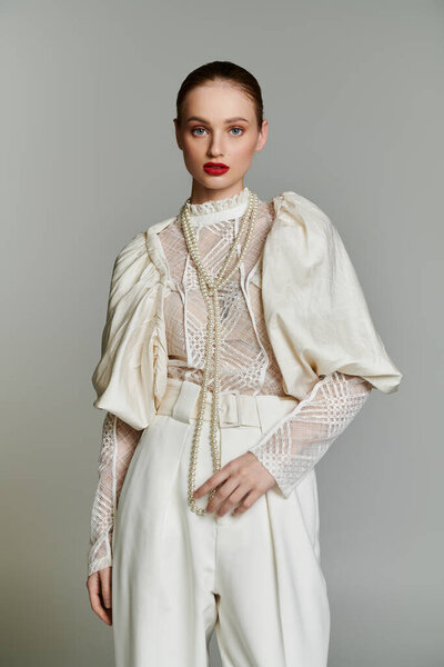 Young woman in white lace blouse, trousers, pearl necklace, serious expression.