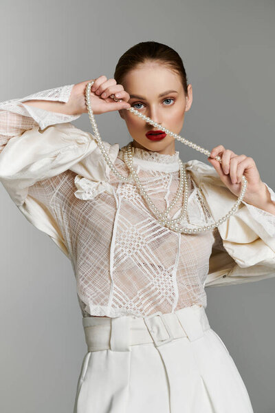 A young woman in a white lace blouse and white pants holds a string of pearls in front of her face.