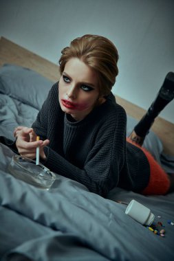 Şık giyinmiş bir kadın yatakta poz verir, elinde sigarayla kameraya bakar..
