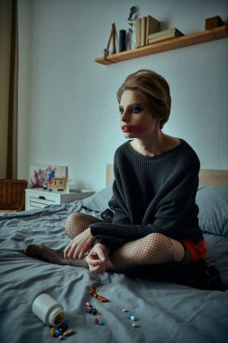 Grunge giysili bir kadın yatakta oturur, yatak örtüsüne yayılmış haplar gibi yansıtır..