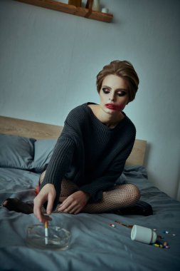 Genç bir kadın yatakta oturuyor, gözleri yerde, etrafa saçılmış haplar ve kül tablası ile çevrili..