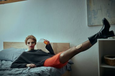 Süslü, grunge kıyafetli bir kadın bacaklarını açmış bir yatakta poz verir..