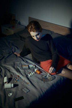 Zarif giyimli bir kadın yatakta oturur, sigara içer ve etrafında haplar ve kül tablası olur..