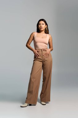 Bronz pantolonlu ve pembe bluzlu bir kadın stüdyo ortamında duruyor..
