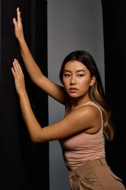 Asyalı bir kadın siyah bir fonda poz veriyor, eli duvarda dinleniyor..