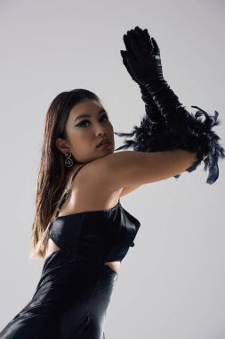 Siyah elbiseli ve tüylü eldivenli bir kadın gri arka planda dramatik bir şekilde poz verir..