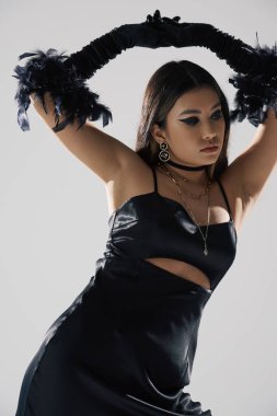 Siyah elbiseli ve tüy eldivenli Asyalı bir kadın beyaz bir fonda poz veriyor..