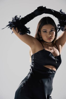 Siyah saten elbiseli, tüy eldivenli bir kadın beyaz bir fonda poz veriyor..