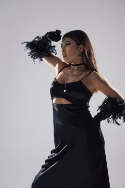 Siyah elbiseli ve tüy eldivenli Asyalı bir kadın beyaz bir arka plana karşı dramatik bir şekilde poz veriyor..