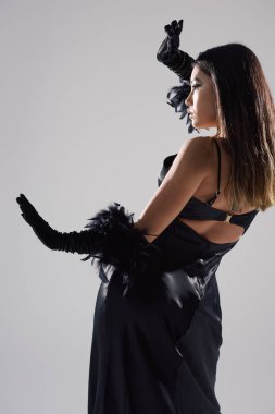 An Asian woman in a black satin dress and feather gloves poses against a light background. clipart