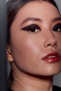 Bir kadının yüzü, gölgeyle kısmen gizlenmiş, dramatik göz kalemi ve kırmızı ruj ile..