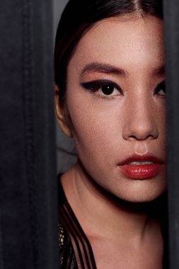 Perdenin arkasındaki kadın yüzü çarpıcı gözleri ve kalın kırmızı ruju gösteriyor..