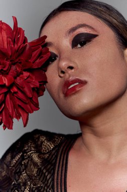 Çarpıcı koyu renk göz kalemi olan bir kadın yoğun bir şekilde bakar, eli kırmızı bir çiçeği kucaklamaktadır..