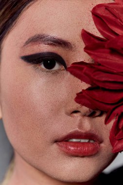Bir kadının yüzü kırmızı bir çiçek tarafından kısmen gizlenir ve dramatik bir göz kalemi ile tek bir göz ortaya çıkar..