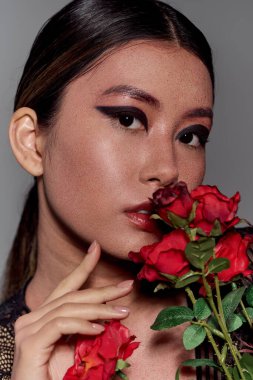 Koyu renk saçlı ve dramatik makyajlı bir kadın elinde kırmızı güllerle poz veriyor..