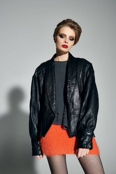 Женщина в кожаной куртке и пушистой оранжевой юбке позирует на белом фоне.