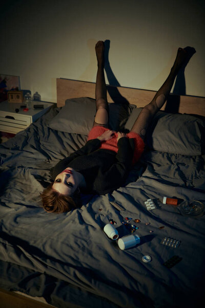 A woman in stylish attire lays on a bed, legs in the air.