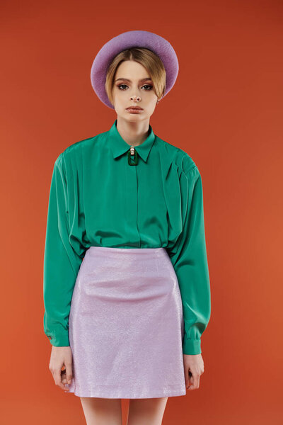 Женщина в лавандовой мини-юбке и зеленой рубашке позирует на оранжевом фоне.