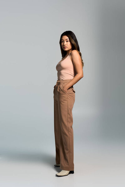 Woman in studio, confidently gazing, wearing pink tank top and brown pants.