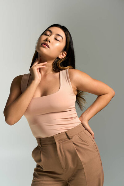 Молодая женщина в розовой майке и коричневых брюках позирует на нейтральном фоне.