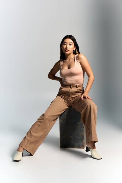 Азиатская женщина в светло-розовом топе и широконогих коричневых штанах позирует на бетонном блоке.