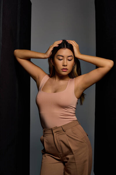 A young woman with long dark hair stands against a gray background, her hands on her head.