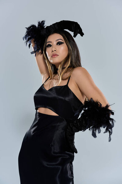 Женщина в гладком черном платье с перьевым акцентом уверенно позирует на белом фоне.