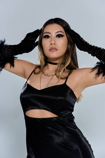 Азиатка в гладком черном атласном платье и перчатках с уверенностью позирует.