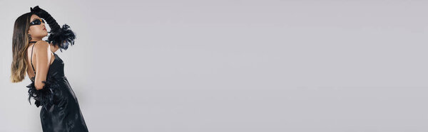 Стильная азиатка в черном платье и перчатках позирует на белом фоне.