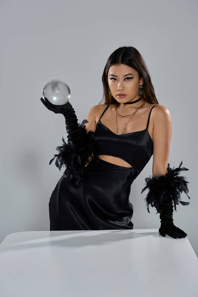 Женщина в гладком черном платье и перчатках пристально смотрит на хрустальный шар.