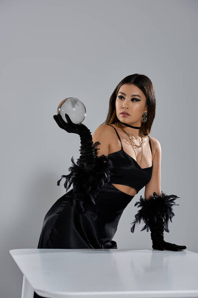 Женщина в черном платье и перьях пристально смотрит сквозь хрустальный шар.