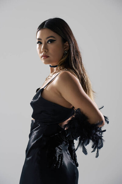 Женщина с длинными темными волосами, одетая в черное платье и перья на перчатках, смотрит через плечо.