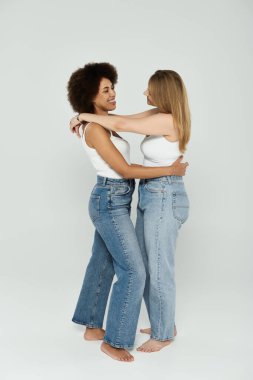 Beyaz atlet giyen ve kot pantolon giyen iki kadın stüdyo ortamında sarılıyorlar..