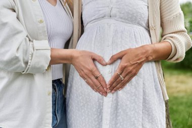 Partneriyle birlikte hamile bir kadın, elleri karnında kalp şekli oluşturuyor..
