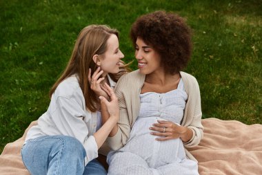 İki kadın, biri hamile, çimenli bir tarlada battaniyede otururken birbirlerine sevgi dolu bakışlar atıyorlar..