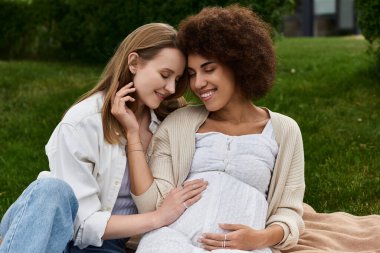 A pregnant woman and her partner share a tender moment in a green grassy field, their hands resting on the growing belly. clipart