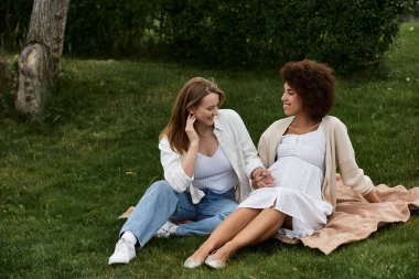 İki kadın, biri hamile, çimenli bir parkta battaniyeye oturmuş, gülümsüyor ve birbirlerine sevgiyle bakıyorlar..