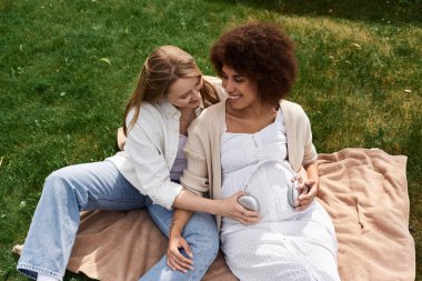 İki kadın, biri hamile, parkta battaniyede müzik dinlerken sarılıp gülümsüyorlar..