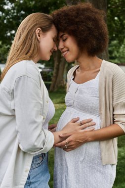 A pregnant Black woman and her partner, a White woman, embrace and touch the woman's belly, radiating joy and anticipation. clipart